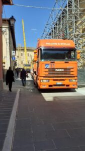 Camion arancione su strada con impalcature e persone.