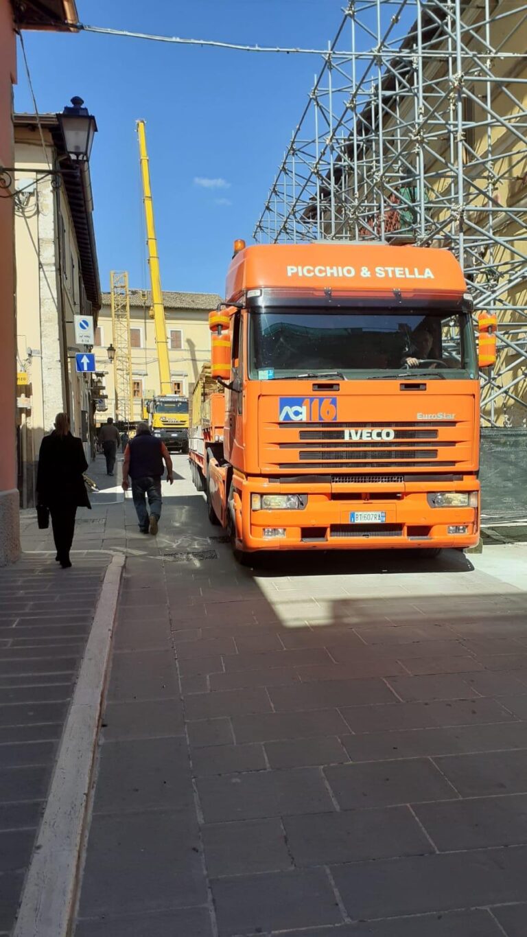 Camion arancione su strada con impalcature e persone.